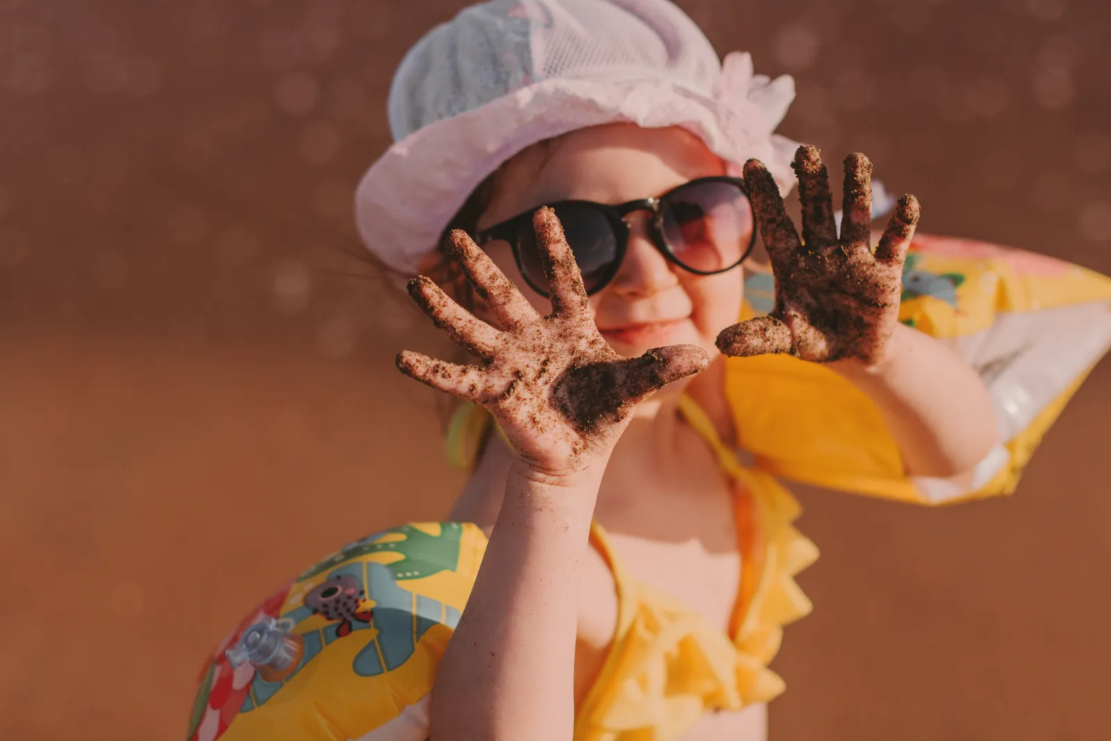 A little girl at the beach holding up her sand-covered hands, wearing sunglasses and a sun hat