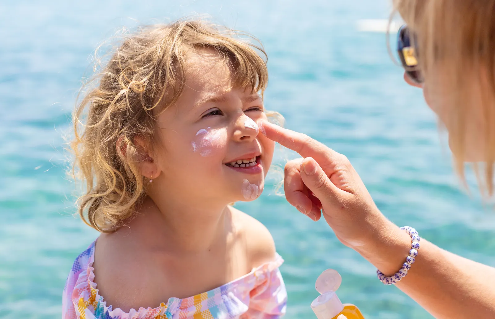A parent applying sunscreen to a smiling child's face and cheeks by the water